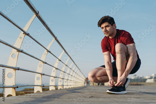 Full size strong sporty athletic toned fit sportsman man in sports clothes sit laces up sneakers shoes warm up train at sunrise sun dawn over sea beach outdoor on pier seaside in summer day morning © ViDi Studio