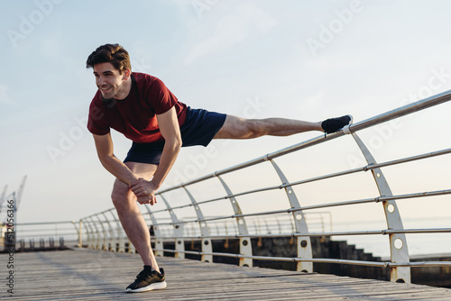 Full length side view young strong sporty fit sportsman man 20s in sports clothes do stretch exercise for legs warm up training at sunrise sun dawn over sea beach outdoor on pier seaside in morning © ViDi Studio