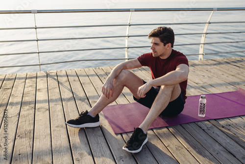 Full size young strong sporty athletic toned fit sportsman man in sports clothes sitting on yoga mat warm up training at sunrise sun dawn over sea beach outdoor on pier seaside in summer day morning