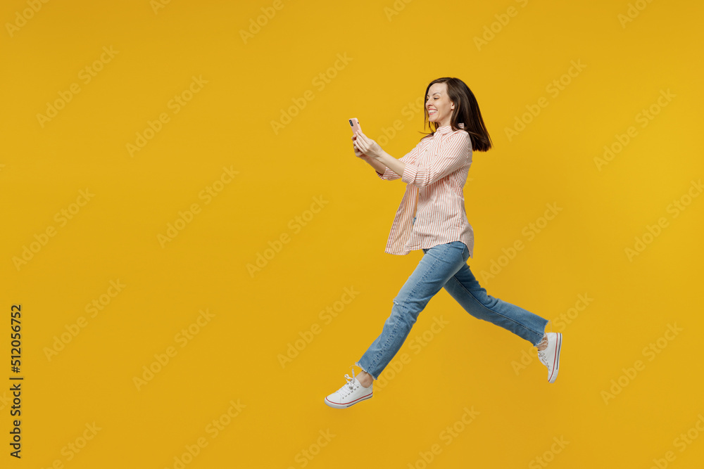 Full body side view young fun woman she 30s in striped shirt white t-shirt jump high run fast hurry up hold use mobile cell phone isolated on plain yellow background studio. People lifestyle concept