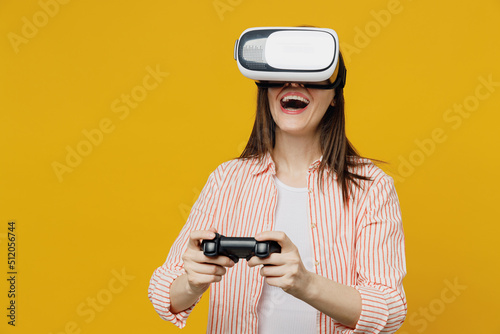 Young happy smiling woman she 30s in striped shirt white t-shirt hold in hand play pc game with joystick console watching in vr headset pc gadget isolated on plain yellow background studio portrait.