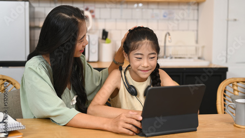Adorable little asian girl doing homework  learning online at home and her mother sitting nearby and giving support