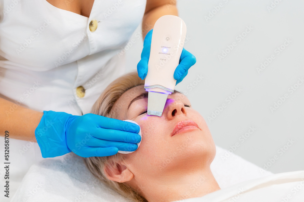 Naklejka premium A professional cosmetologist in blue medical gloves does an ultrasound facial cleansing for an adult Caucasian woman. Hardware anti-aging cosmetology