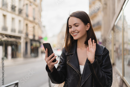 pleased young woman in stylish jacket waving hand while having video chat on street in paris.