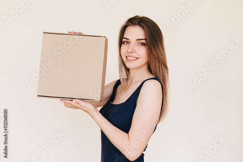 Young Slender Woman in Black Dress on Beige Background With Cardboard Box in Her Hands Closeup. Copy space. Natural Looking Girl Promotes Delivery