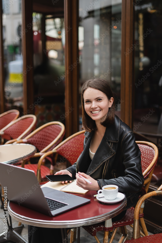 happy freelancer in black jacket holding smartphone near laptop and cup of coffee in french outdoor cafe.