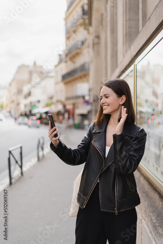 happy young woman in stylish jacket waving hand while having video chat on street in paris.