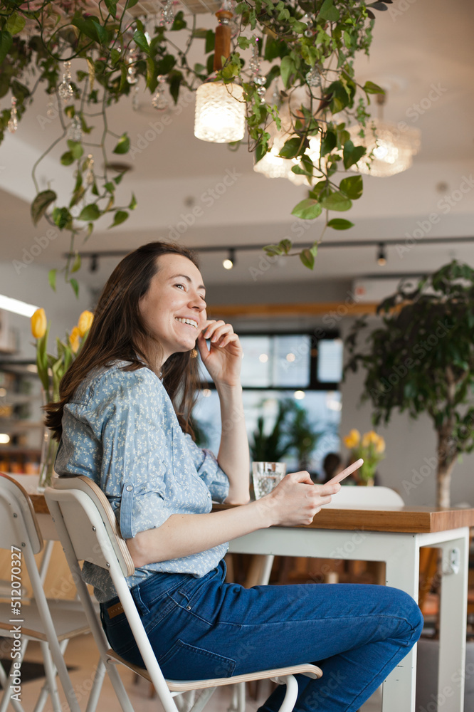 Young beautiful woman using mobile phone to browse social media sitting at cafe or coworkring space, smiling. Candid lifestyle photo of female using technology Young beautiful woman using mobile phone to browse social media sitting at cafe or coworkring space, smiling. Candid lifestyle photo of female using technology