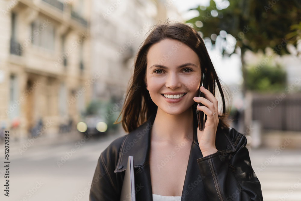 portrait of happy woman in black leather jacket holding laptop and talking on smartphone on street in paris. portrait of happy woman in black leather jacket holding laptop and talking on smartphone on street in paris.