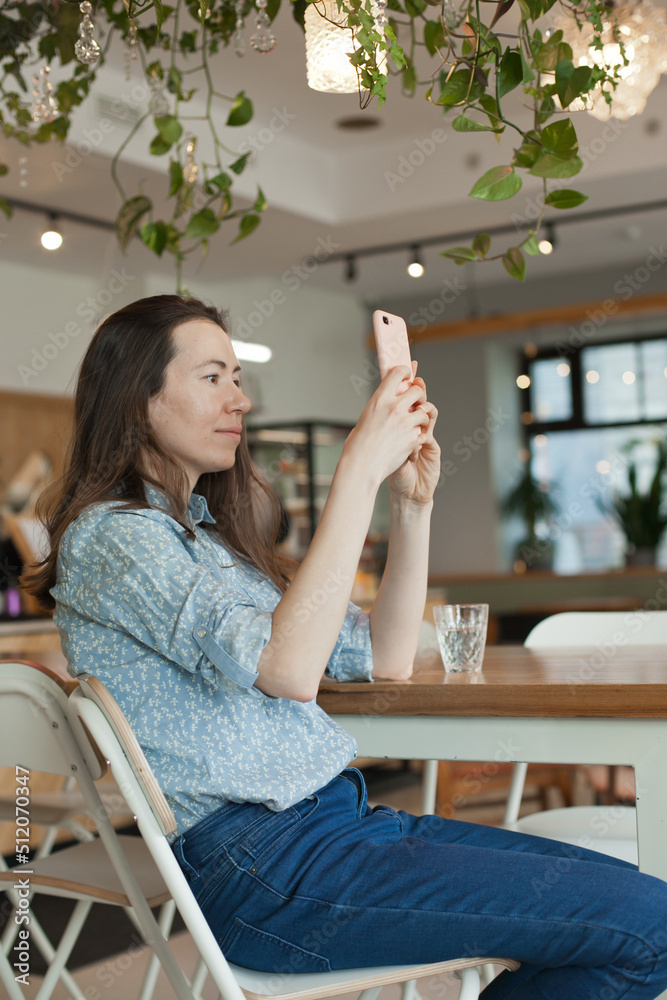 Young beautiful woman using mobile phone to produce content for social media making video or photo sitting at cafe or coworkring space, smiling. Candid lifestyle photo of female using technology Young beautiful woman using mobile phone to produce content for social media making video or photo sitting at cafe or coworkring space, smiling. Candid lifestyle photo of female using technology