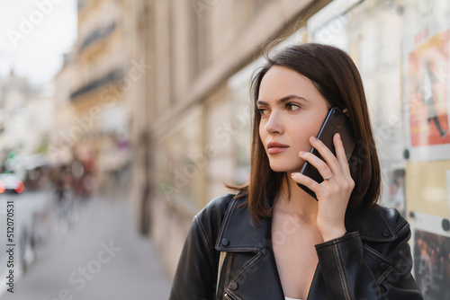 portrait of brunette young woman in stylish jacket talking on smartphone on street in paris.