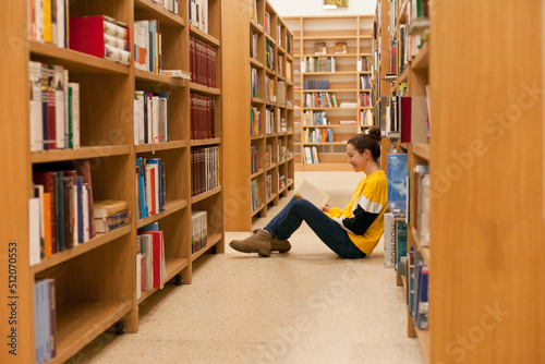 Young female student sitting on the floor near bookshelf reading a book. Woman wearing jeans and yellow sweatshirt studying while sitting in college library or bookstore. 