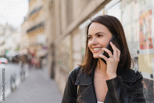 portrait of joyful young woman in stylish jacket talking on smartphone on street in paris.