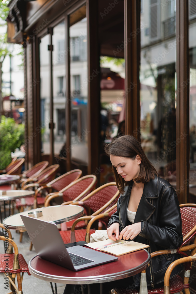 stylish freelancer in black leather jacket looking at notebook while sitting near laptop in french outdoor cafe. stylish freelancer in black leather jacket looking at notebook while sitting near laptop in french outdoor cafe.