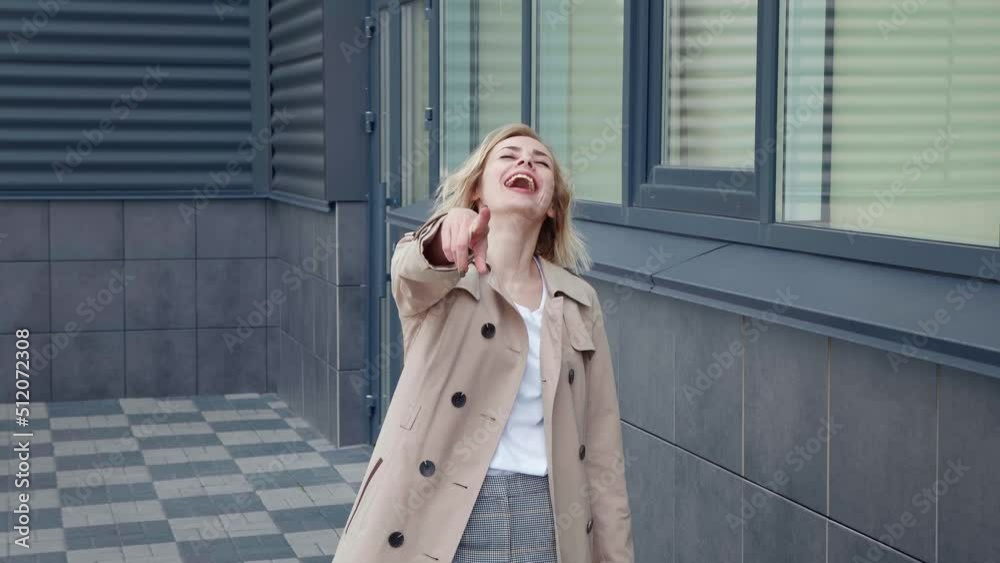 Portrait of laughing young woman pointing index finger into camera, standing on modern office building background. Funny female in raincoat smiling outdoors in slow motion. True emotions concept Portrait of laughing young woman pointing index finger into camera, standing on modern office building background. Funny female in raincoat smiling outdoors in slow motion. True emotions concept