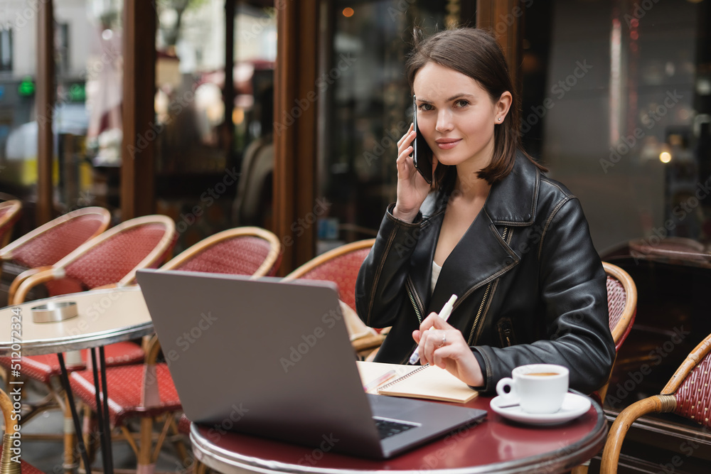 young freelancer in black jacket talking on smartphone near laptop and cup of coffee on table in outdoor cafe in paris. young freelancer in black jacket talking on smartphone near laptop and cup of coffee on table in outdoor cafe in paris.