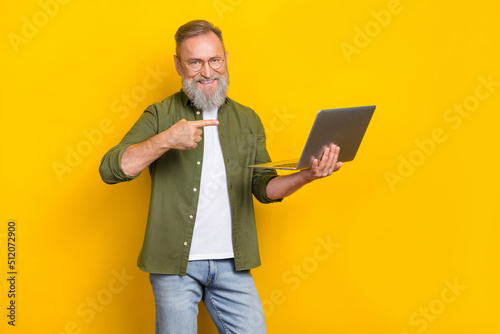 Portrait of cheerful aged man direct finger arm hold wireless netbook isolated on yellow color background