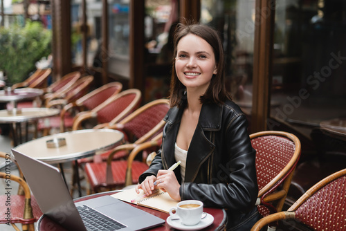 happy woman in black jacket looking at camera near laptop and cup of coffee on table in french outdoor cafe.