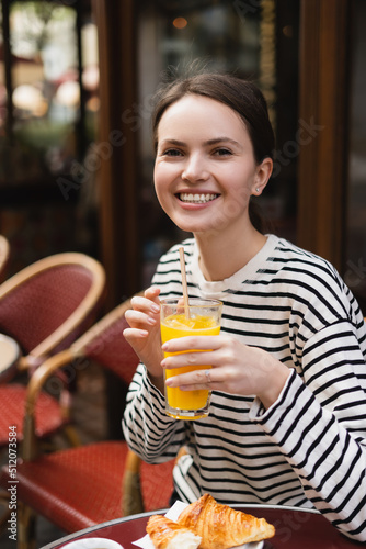 joyful young woman in striped long sleeve shirt holding glass of fresh orange juice in outdoor cafe in paris.