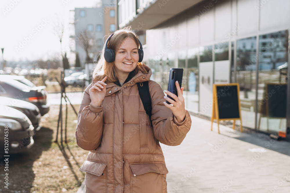 Fototapeta premium Portrait of young happy woman walking on the street in the city. Girl talking on a video call with phone and headphones outdoor. Young woman listening music in headphones in the city.