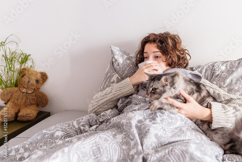 young woman lies in bed in warm clothes, covered with blanket, blows nose, fluffy gray domestic cat lies nearby. concept of allergic rhinitis on animal fur, viral infection, cold, flu, runny nose photo