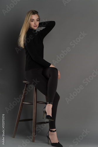 A young attractive girl in black tight-fitting clothes and black high-heeled shoes is sitting on a high stool. Full height