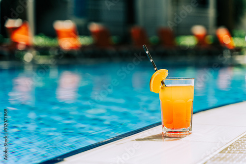 Tropical sparkling cocktail by the pool. The picture of glass with orange lemonade fruit cocktail standing near the poolside. Summer alcohol free drink by the hotel pool. Hello summer holiday vacation