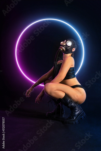 Seductive girl fetish model with a slim figure poses sitting in a black gas mask, black leather or latex sexy underwear and leather boots with neon lights on the background. bdsm, fetish, polluted air