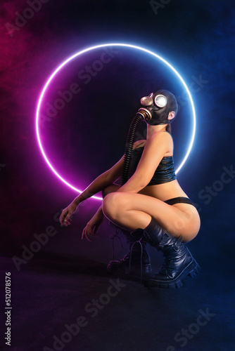 Seductive girl fetish model with a slim figure poses sitting in a black gas mask, black leather or latex sexy underwear and leather boots with neon lights on the background. bdsm, fetish, polluted air