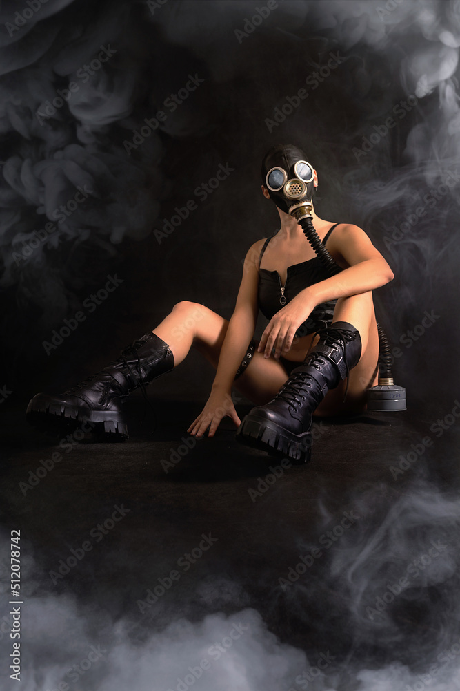Seductive girl fetish model with a slim figure poses sitting in a black gas mask, black leather or latex sexy underwear and leather boots on a black background. BDSM, perversion, fetish, smoke around