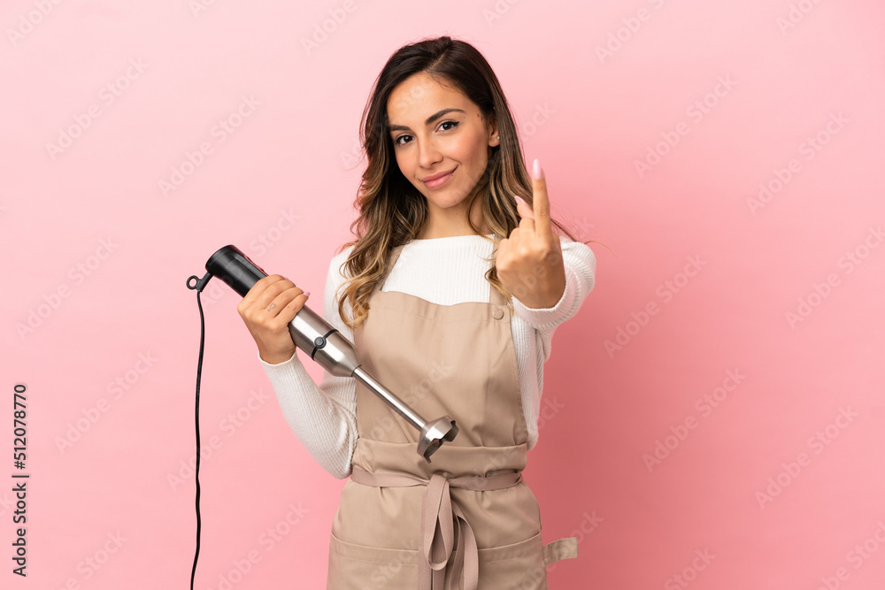 Young woman using hand blender over isolated pink background doing coming gesture