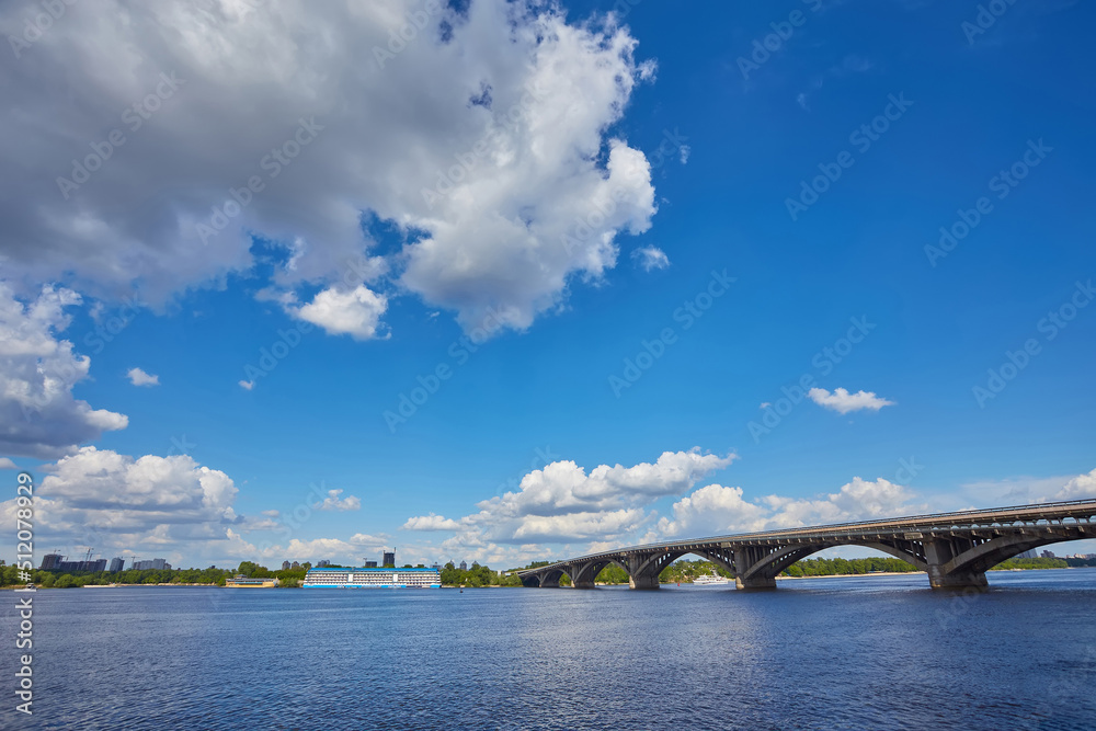 View from the right bank of the Dnieper river over the first metro bridge in Kyiv city with blue subway train, motorboat under it and hotelship behind.