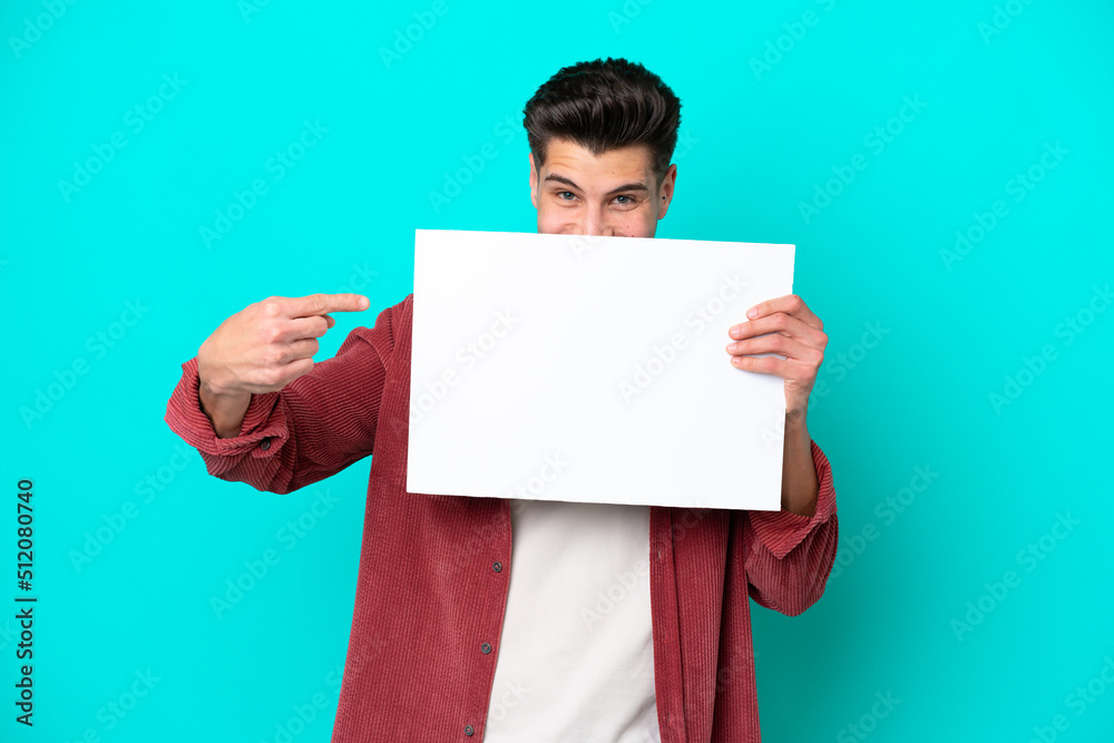 Young handsome caucasian man isolated on blue bakcground holding an empty placard and hiding behind it Young handsome caucasian man isolated on blue bakcground holding an empty placard and hiding behind it