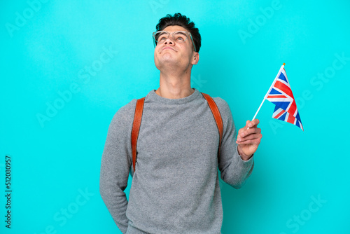 Young caucasian man holding an United Kingdom flag isolated on blue background and looking up