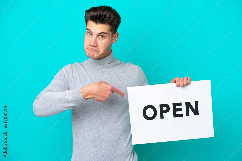 Young handsome caucasian man isolated on blue bakcground holding a placard with text OPEN and pointing it Young handsome caucasian man isolated on blue bakcground holding a placard with text OPEN and pointing it