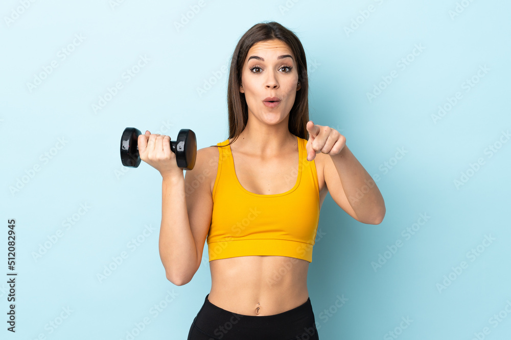 Young caucasian woman making weightlifting isolated on blue background surprised and pointing front Young caucasian woman making weightlifting isolated on blue background surprised and pointing front