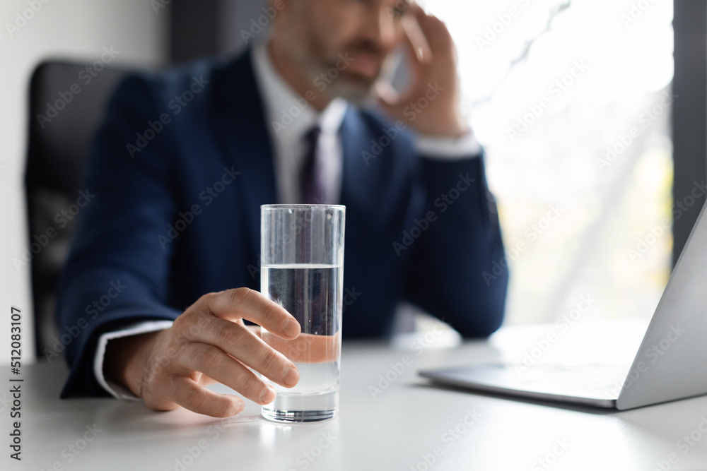 Stressed businessman suffering from headache at workplace, holding glass of water Stressed businessman suffering from headache at workplace, holding glass of water