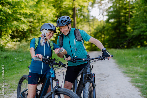 Portrait of active senior couple riding bicycles at summer park, looking at sports smartwatch, checking their performance.