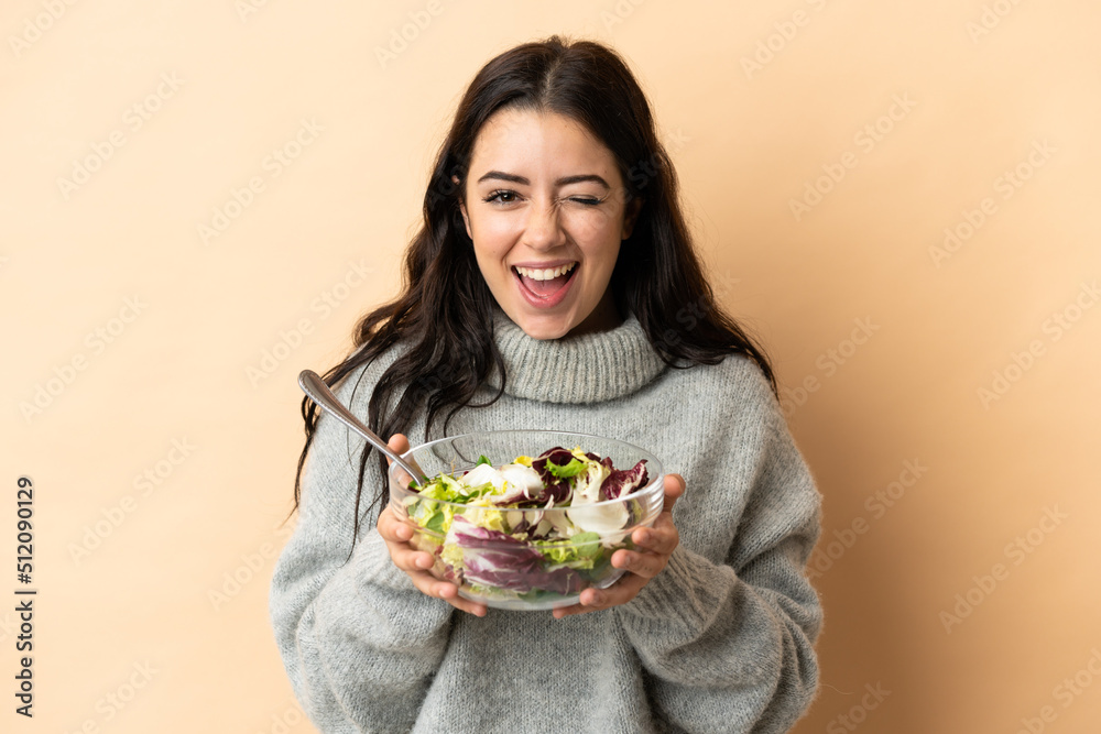 Young caucasian woman isolated on beige background holding a bowl of salad while winking Young caucasian woman isolated on beige background holding a bowl of salad while winking