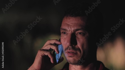 Concerned young man speaks on the phone at night in the forest. Man in the dark, illuminated face. Background out of focus with backlight. Nightlife, summer outdoors. 4K UHD.