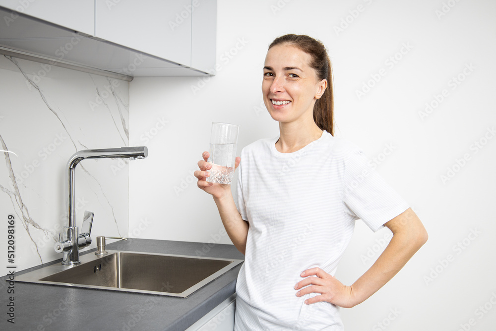 Smiling young woman in a white t-shirt in the kitchen drinks water from a glass cup Smiling young woman in a white t-shirt in the kitchen drinks water from a glass cup