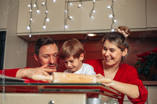 Cute young family spends time in kitchen at home, dressed in red, baking Christmas cookies. Laughing, having fun. Joyful evening for dad, mom and son.