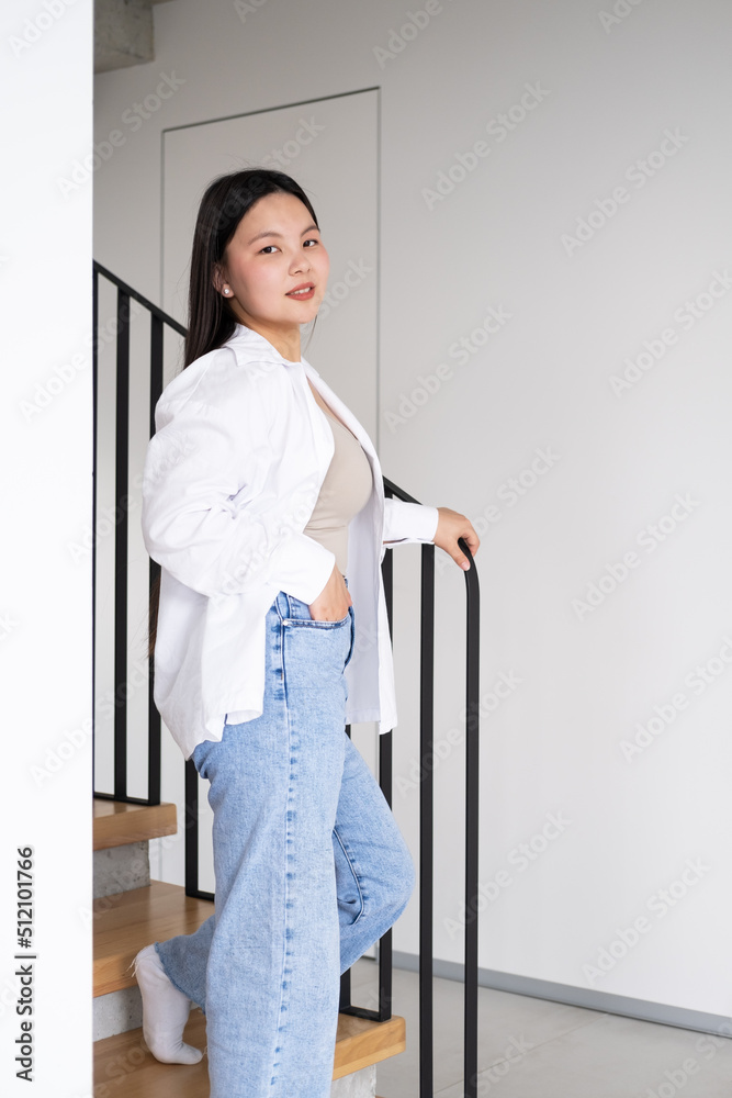 beautiful smiling young asian woman with long dark hair in white shirt on stairs at modern interior beautiful smiling young asian woman with long dark hair in white shirt on stairs at modern interior