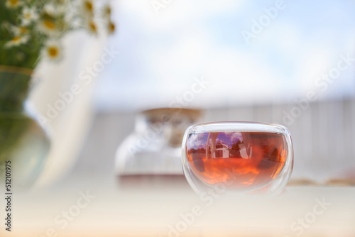 Transparent glass Cup of tea in nature. The concept of breakfast in the backyard of the house. Early morning, tea and kettle. out of focus, chamomile flowers in the background and blue sky