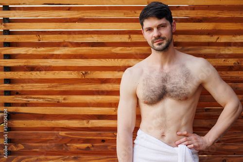 Strong confident man with attractive body in a towel posing against the background of a wall of wooden planks on a spa resort