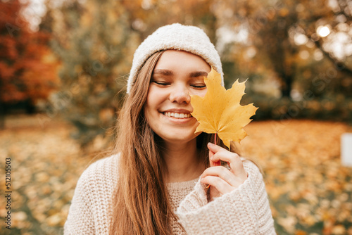 Charming young woman in knitted hat and sweater smiles happily on walk in autumn park  holding yellow maple leaf in front of her. Enjoying autumn nature. Leaf fall. Positive emotions.