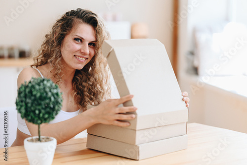 Cardboard Boxes Are Stacked On Wooden Table In Kitchen. Natural Light from the Window. Young woman opens the top box and happily looks into camera.