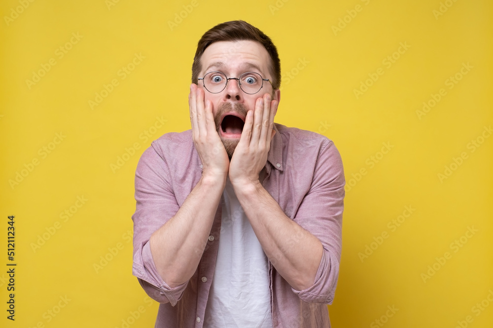 Man with glasses is shocked and frightened, he screams with mouth wide open, holds hands on cheeks and looks with big eyes. Man with glasses is shocked and frightened, he screams with mouth wide open, holds hands on cheeks and looks with big eyes.