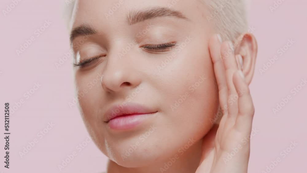 Pretty young slim light-haired Caucasian woman strokes her face looking at the camera and smiling for it on pale pink background | Face hydration concept Pretty young slim light-haired Caucasian woman strokes her face looking at the camera and smiling for it on pale pink background | Face hydration concept