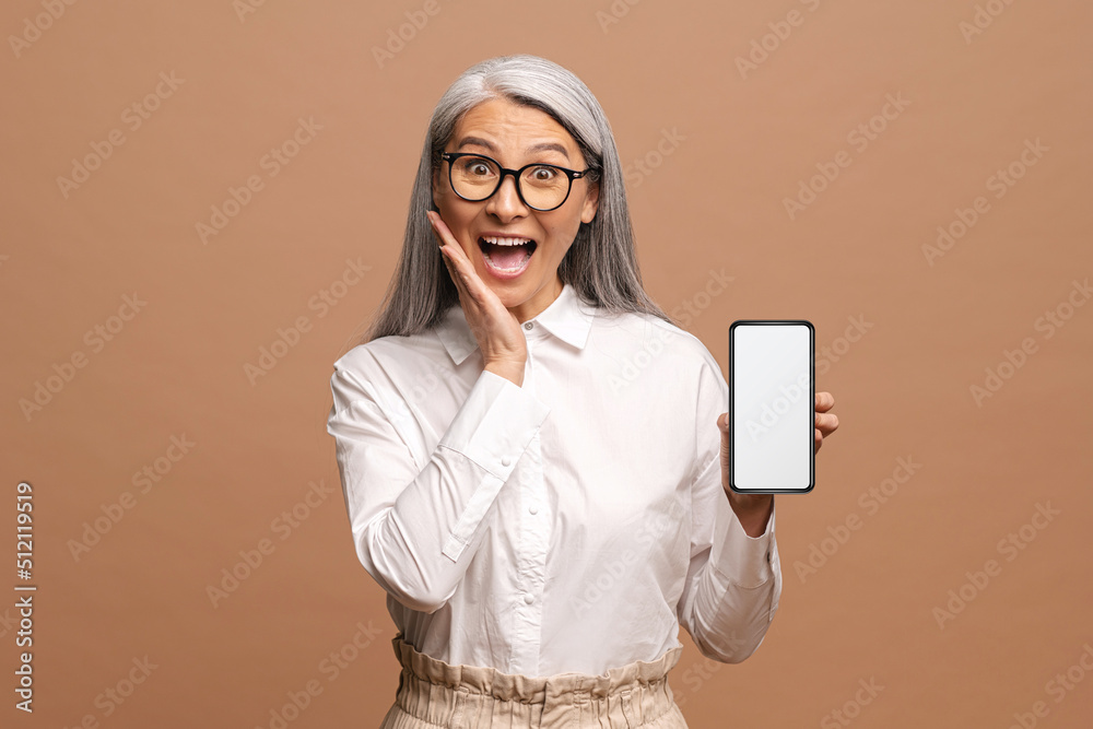 Positive elderly woman looking at the camera and rejoicing with smartphone at her hands isolated over beige background. Advertises or recommends a new product, holiday sale concept Positive elderly woman looking at the camera and rejoicing with smartphone at her hands isolated over beige background. Advertises or recommends a new product, holiday sale concept